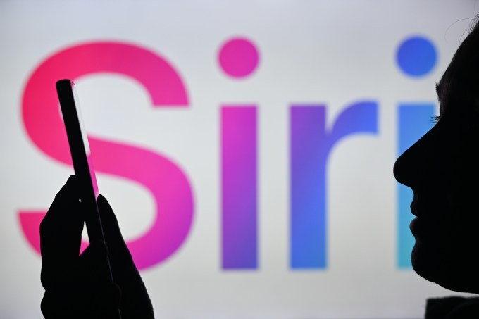 apple-siri-header-1 An image of a woman holding a cell phone in front of the Siri logo displayed on a computer screen.