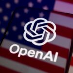 The OpenAI logo appears on the screen of a smartphone placed on a reflective surface onto which the U.S. flag is projected.