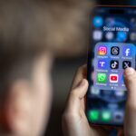 a 14-year-old boy holds an iPhone screen displaying various social media and messaging apps