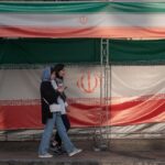 Two young Iranian women walk past the country's flags in northern Tehran, Iran, on June 30, 2025, following the ceasefire between Iran and Israel.