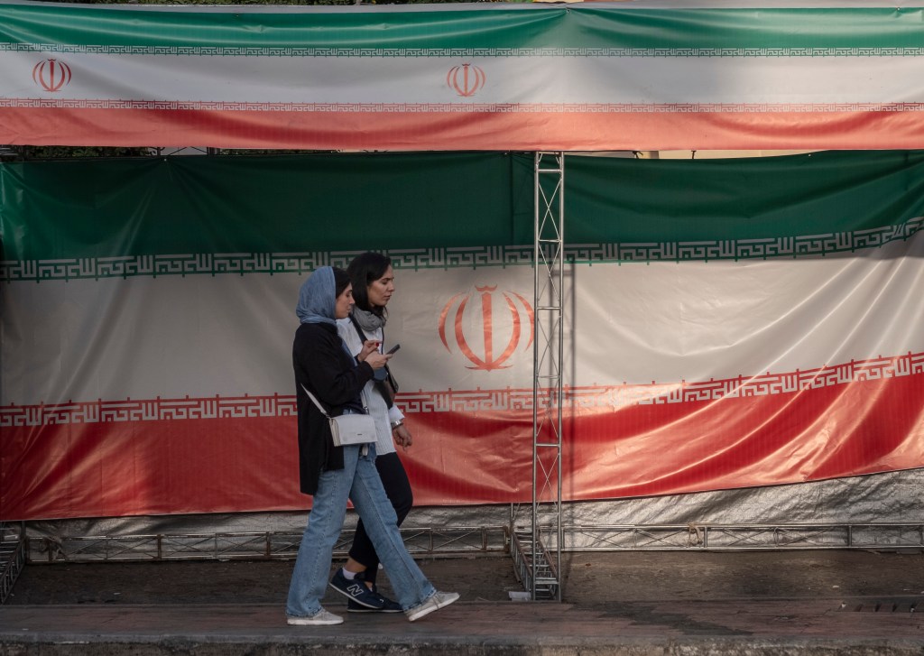 Two young Iranian women walk past the country's flags in northern Tehran, Iran, on June 30, 2025, following the ceasefire between Iran and Israel.