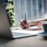 person writing in notebook with laptop open