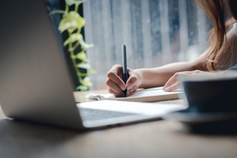 person writing in notebook with laptop open